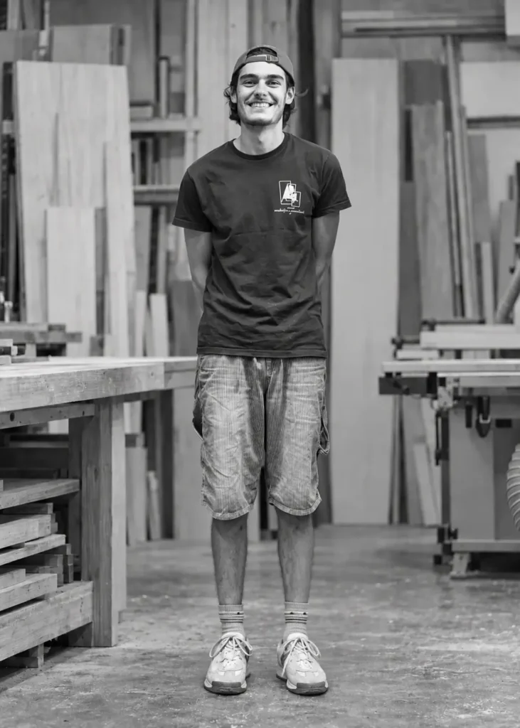 Portrait en noir et blanc d'un ébéniste souriant debout, mains dans les poches, dans son atelier de fabrication de mobilier sur mesure dans le Var.